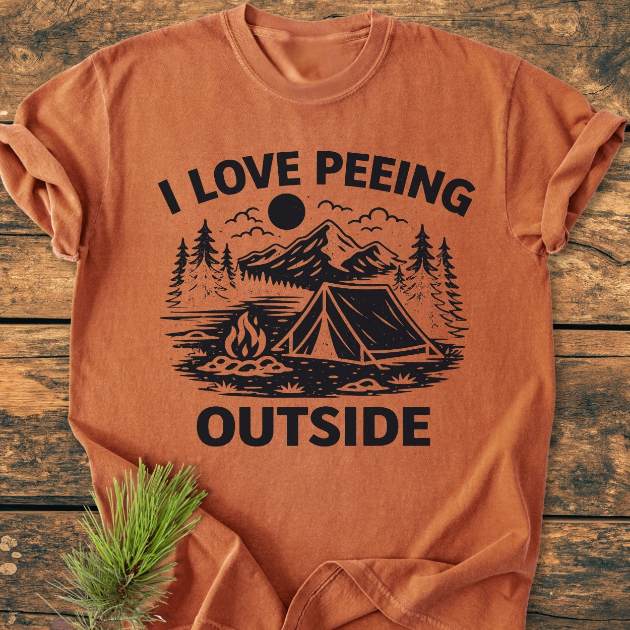 Rust-orange I Love Peeing Outside - Vintage Wash Tee on wood surface, featuring a black campsite graphic with a tent and campfire, accompanied by a small pine sprig.