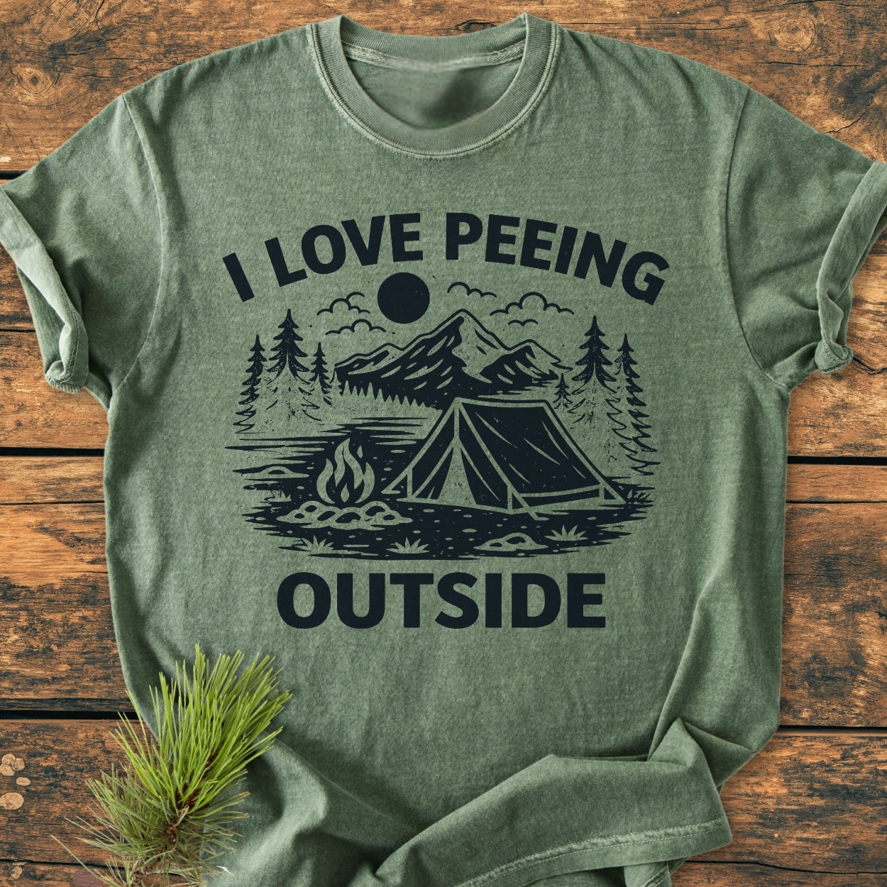 I Love Peeing Outside - Vintage Wash Tee features a heather-green color with a playful camping graphic, showcasing a tent, campfire, and nature elements, laid flat on a wooden surface.