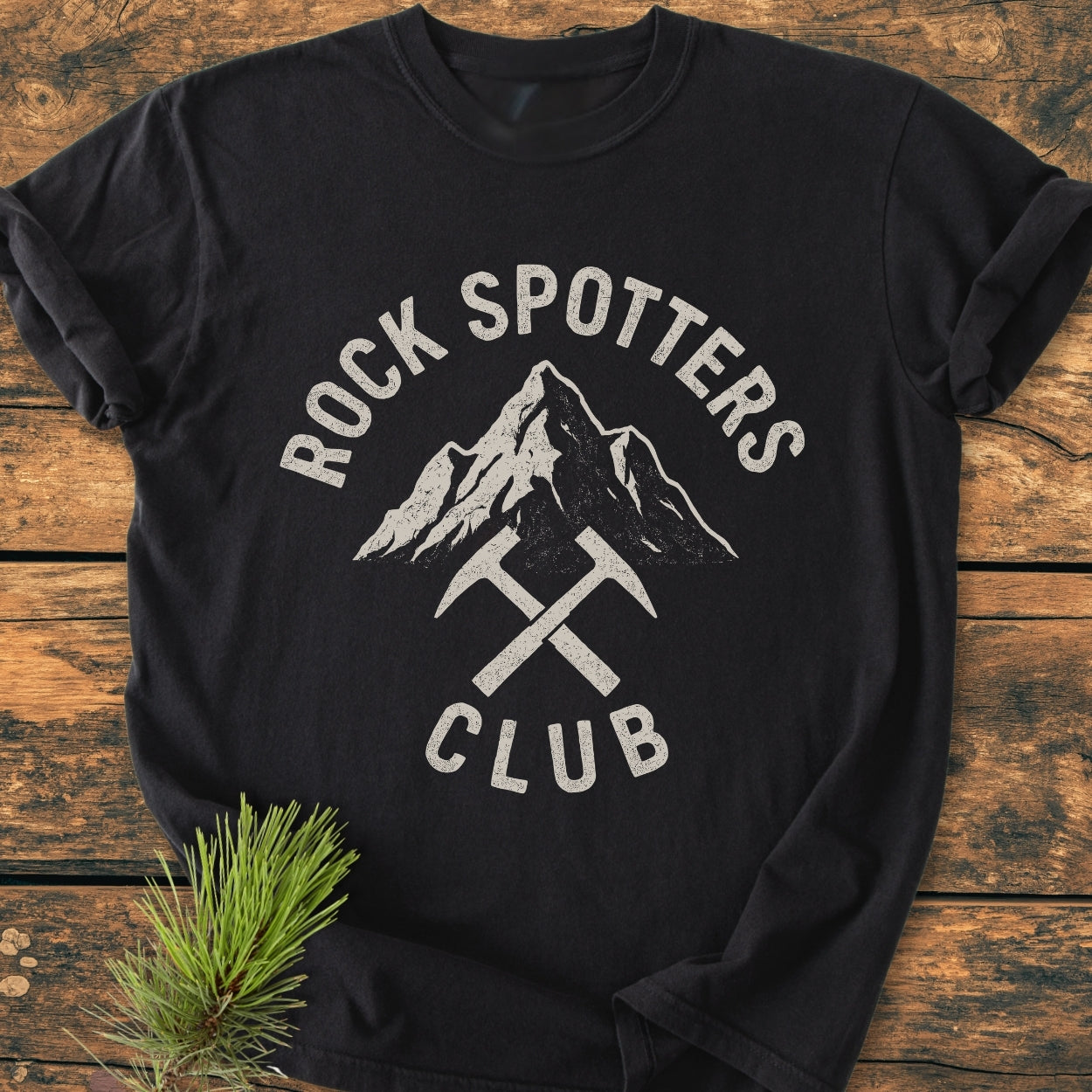 Rock Spotters Club - Vintage Wash Tee featuring a mountain graphic with crossed pick-axes, laid flat on a wooden surface, showcasing its relaxed fit and durable, soft-washed 100% ring-spun cotton fabric.