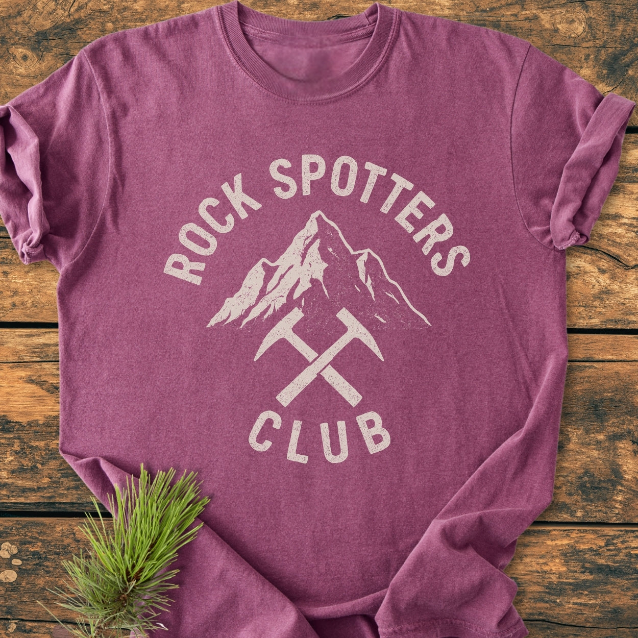 Rock Spotters Club - Vintage Wash Tee in heathered-purple, featuring a white distressed mountain and rock-hammers graphic, laid flat on wood with a pine sprig accentuating its rustic charm.