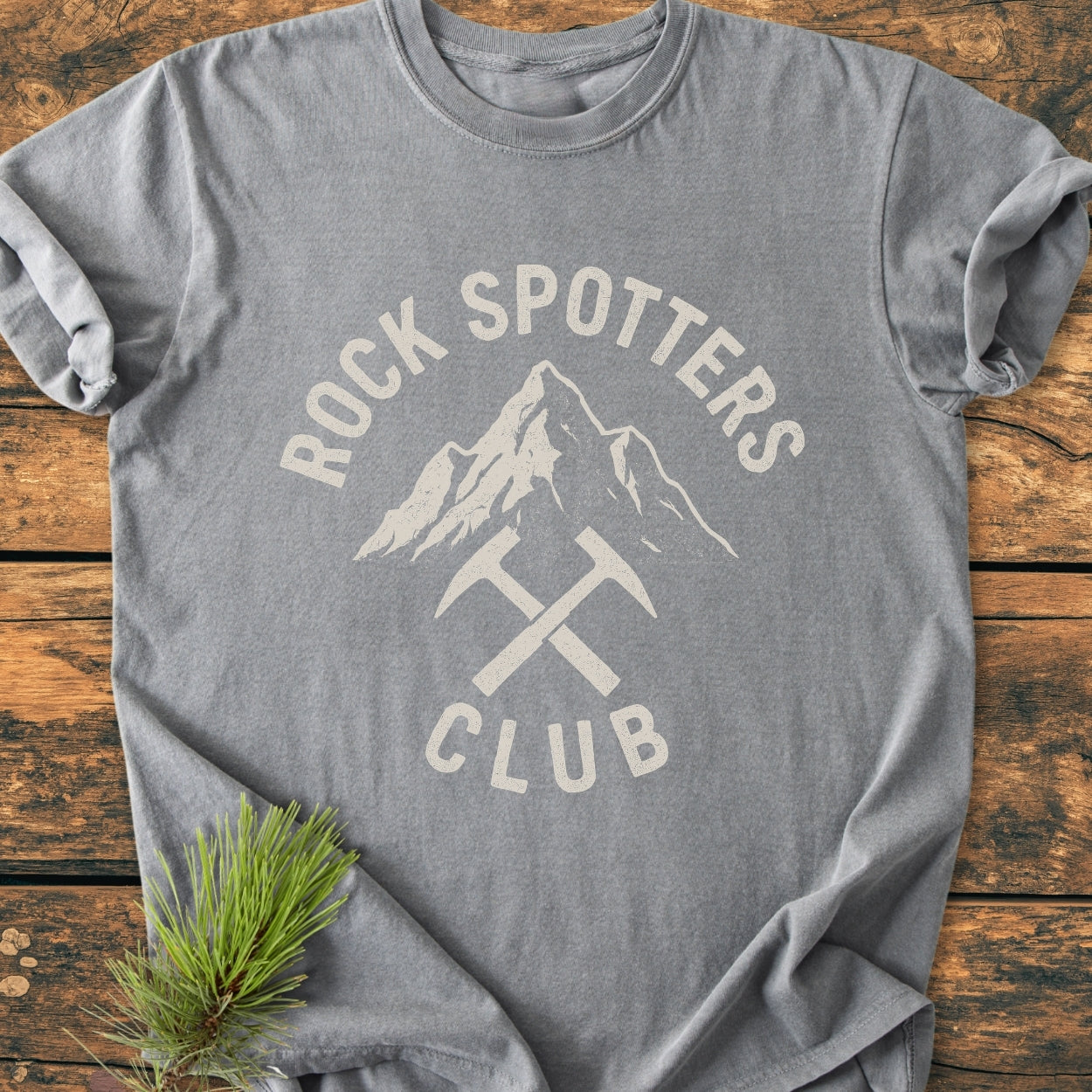 Rock Spotters Club - Vintage Wash Tee features a heather-gray design with a stylized mountain and rock-hammers print, laid on a wooden surface, complemented by a pine sprig.