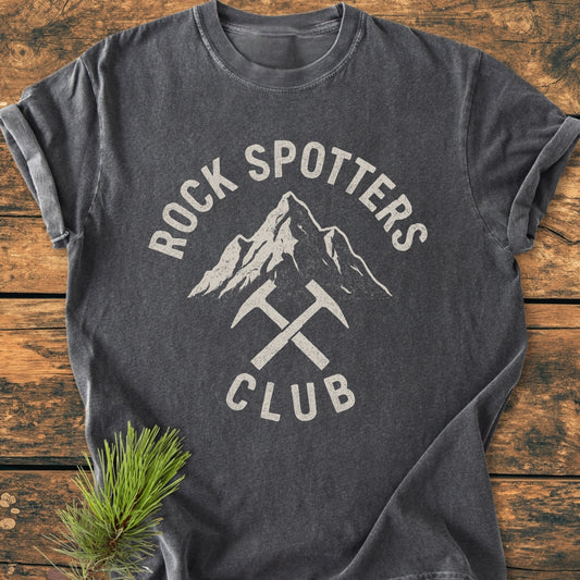 Rock Spotters Club - Vintage Wash Tee features a mountain design with crossed hammers, laid flat on wood, alongside a pine sprig. Made from 100% ring-spun cotton for comfort and durability.