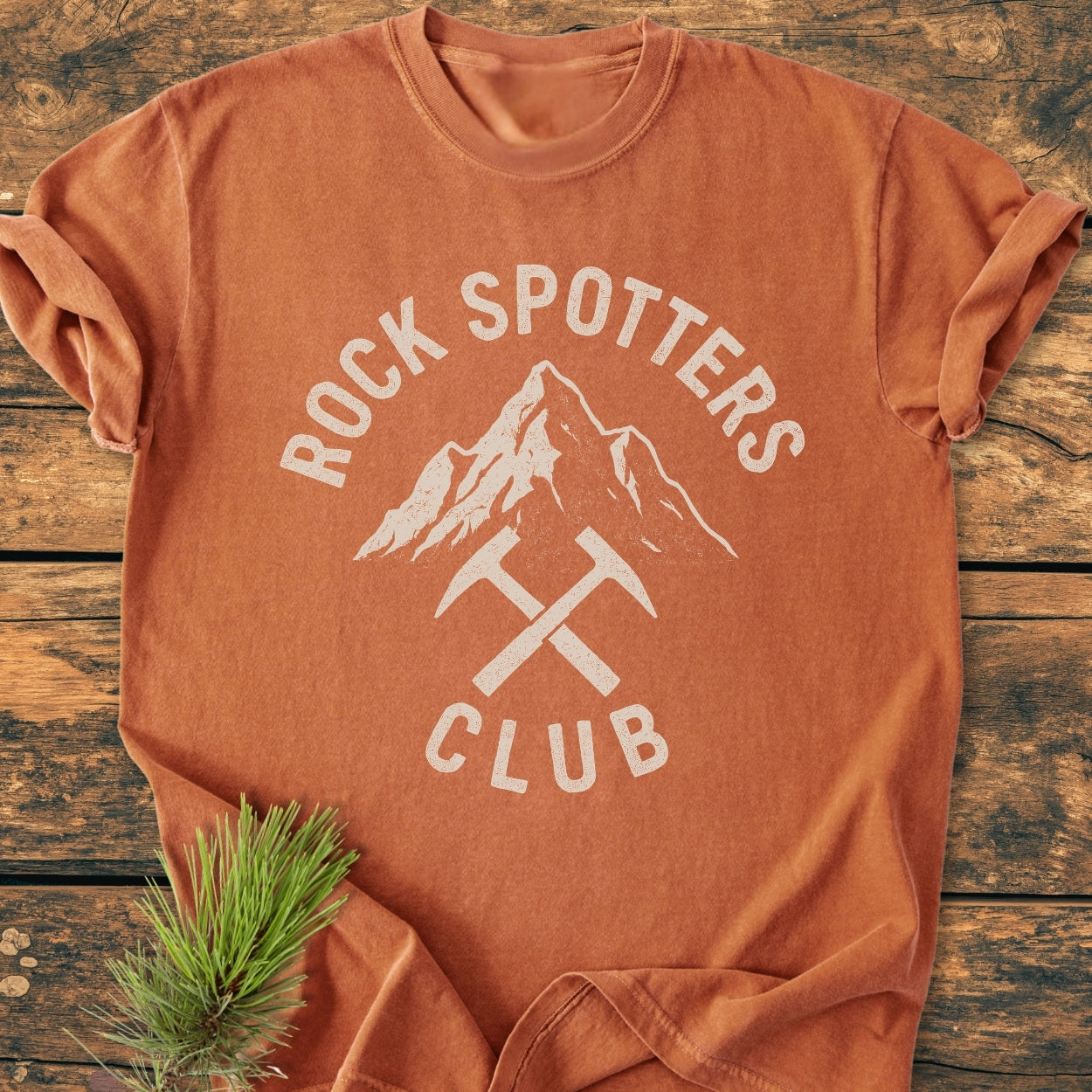 Rock Spotters Club - Vintage Wash Tee featuring an arched logo and mountain graphic design on a burnt-orange shirt, laid on rustic wood with a pine sprig.
