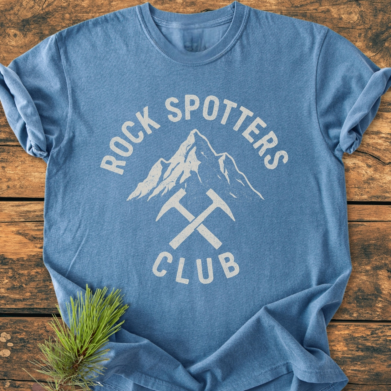 Rock Spotters Club - Vintage Wash Tee displaying a mountain peak and crossed rock-hammers design, laid on wood with a pine sprig. Heather-blue, relaxed fit, garment-dyed for comfort and durability.