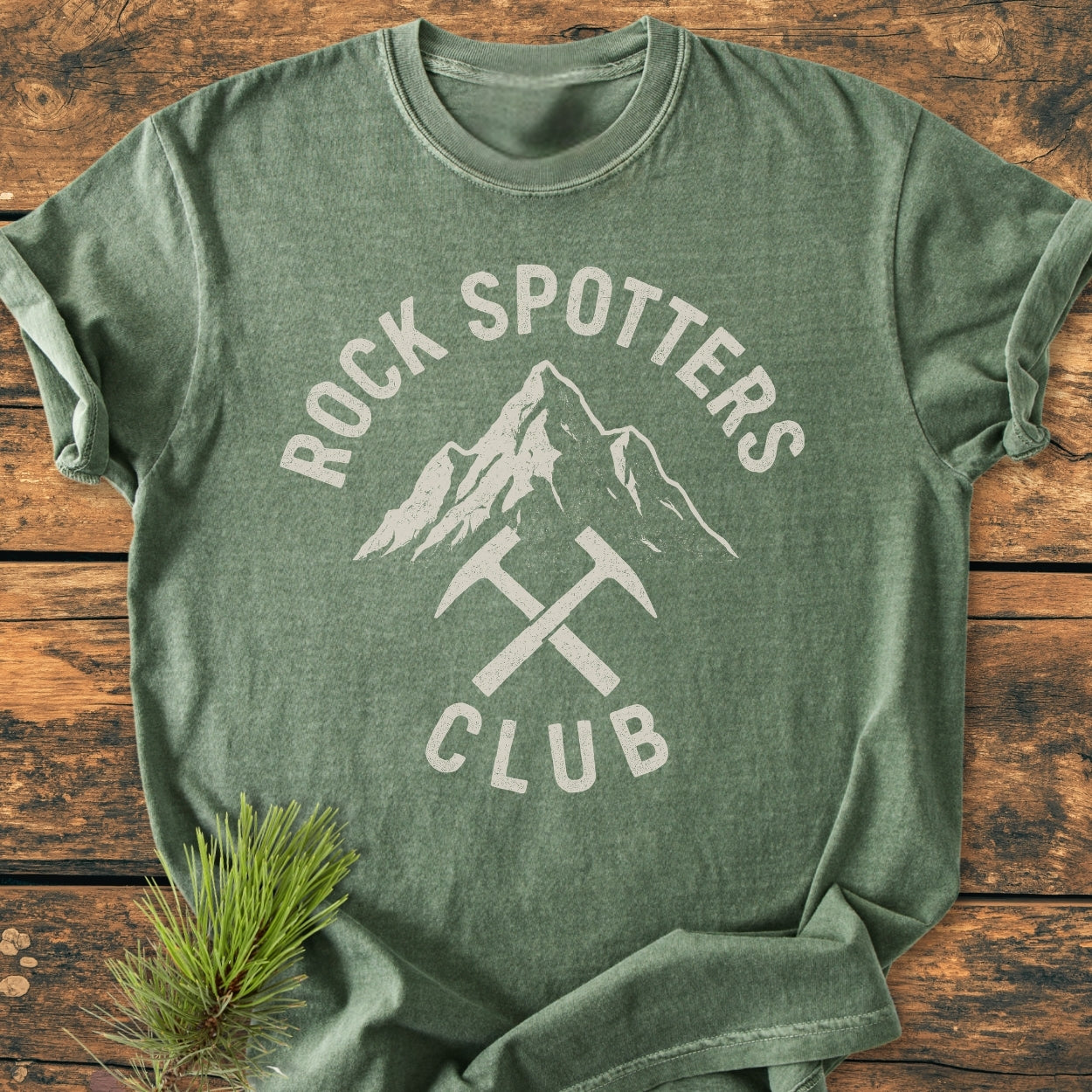Rock Spotters Club - Vintage Wash Tee features a forest-green shirt with a mountain graphic and crossed hammers, laid on wood, accented by a pine branch, highlighting its nature-inspired design.