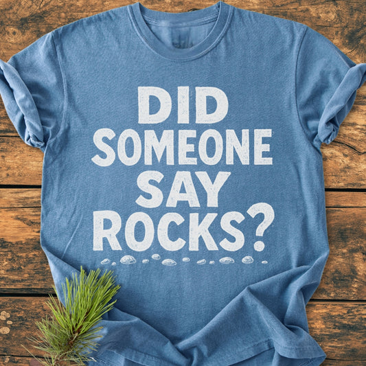 Steel-blue crew-neck tee, Did Someone Say Rocks? in chalk-style white font, rock doodles below, laid flat on wood. Includes a small pine twig detail.