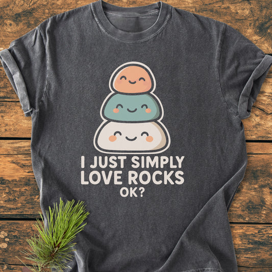 Heather-gray vintage wash tee featuring a graphic of smiling stacked rocks and text I Just Simply Love Rocks OK?, laid on rustic wood with a pine sprig accentuating the natural theme.
