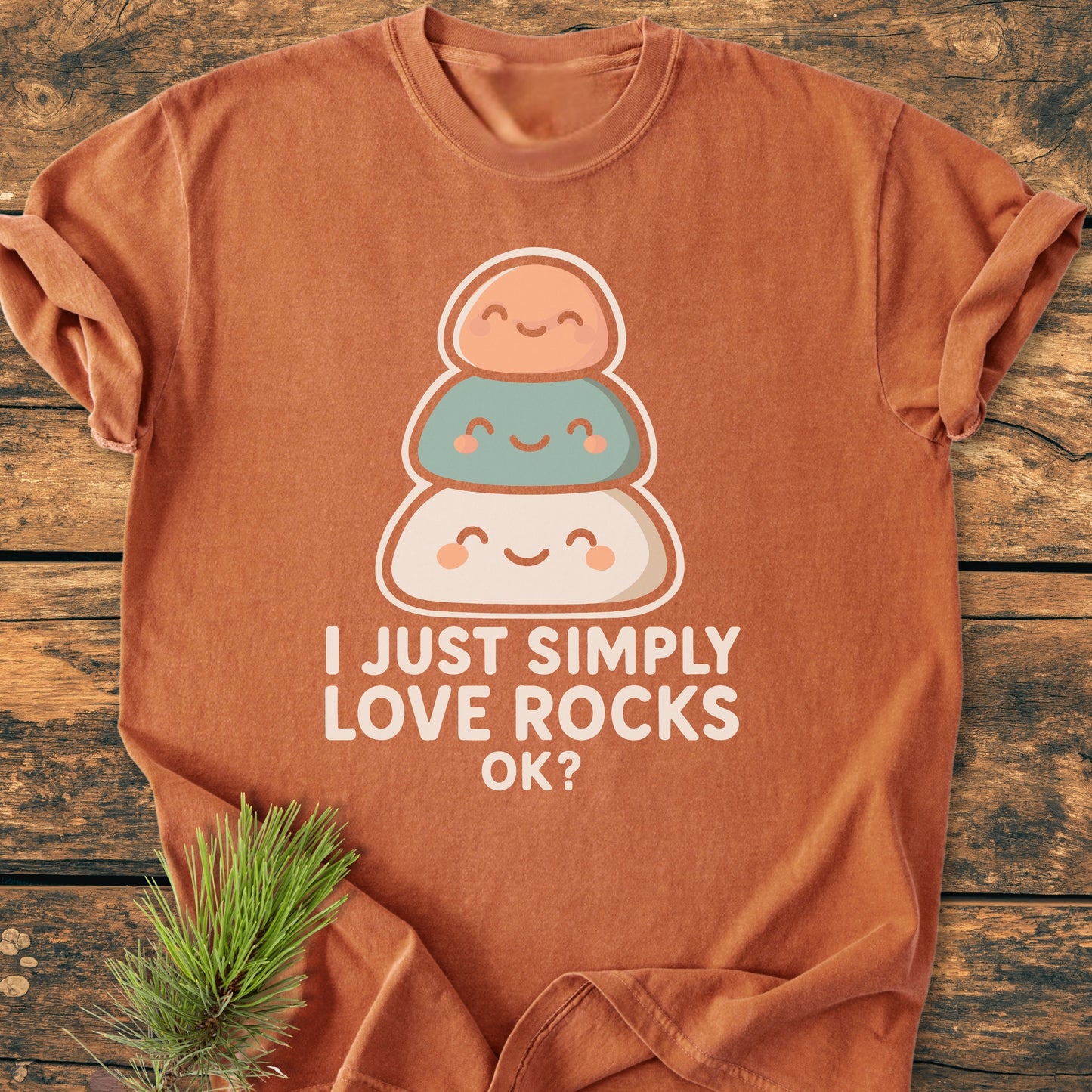 I Just Simply Love Rocks OK? - Vintage Wash Tee features a cartoon of smiling stacked stones on a soft, relaxed fit burnt-orange shirt laid on a wooden surface with pine sprig.