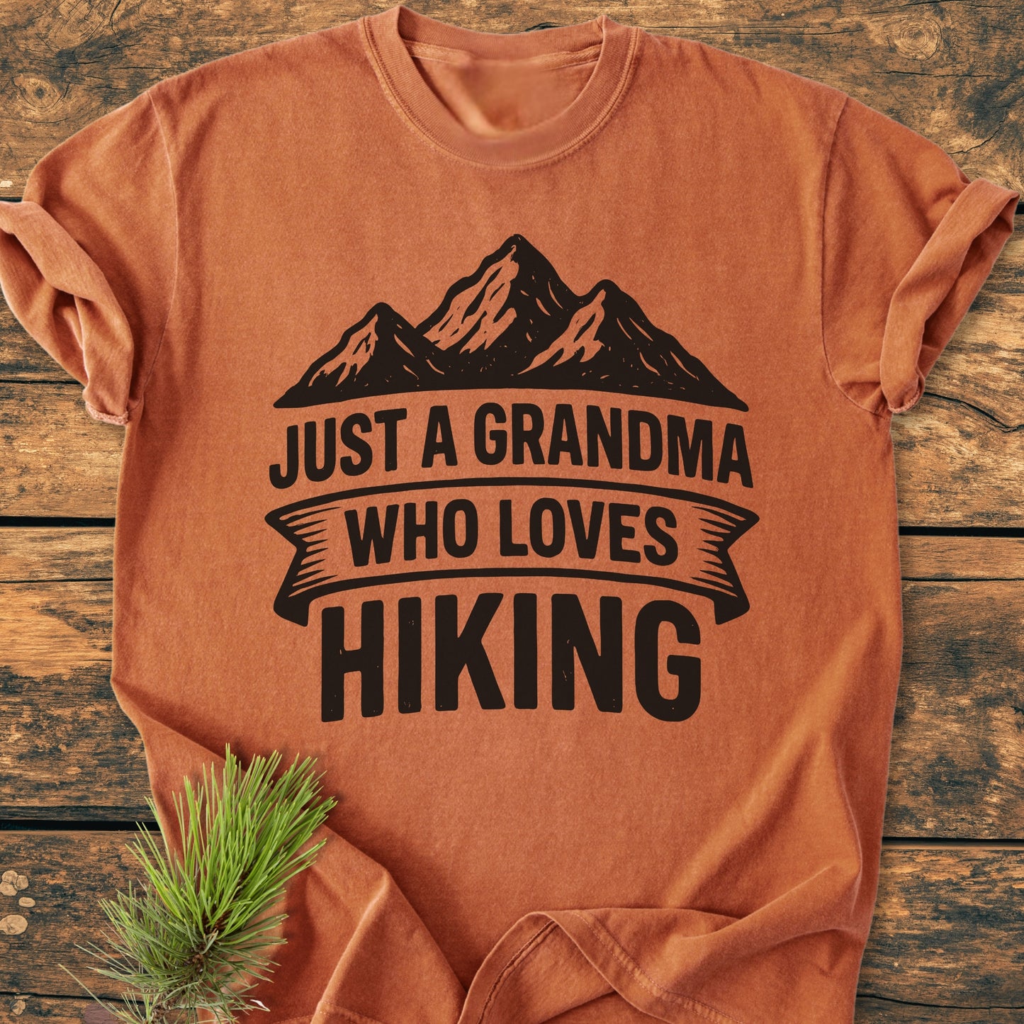 Rust-orange tee, Just A Grandma Who Loves Hiking - Vintage Wash Tee, features mountain graphic and slogan. Laid flat on wood, soft-washed fabric, relaxed fit, durable double-needle stitching, 100% ring-spun cotton.