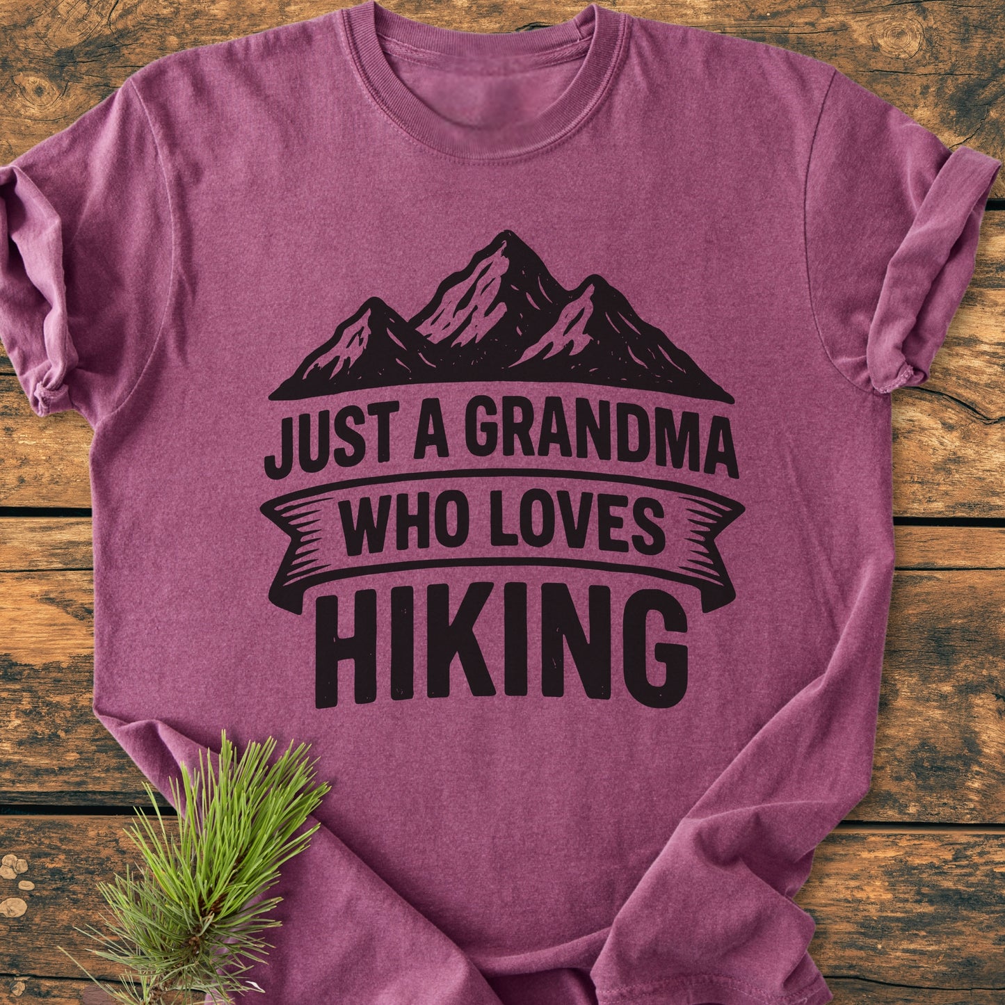 Just A Grandma Who Loves Hiking - Vintage Wash Tee featuring a bold mountain graphic and text, displayed on a rustic surface with a pine sprig accentuating its nature-inspired theme.