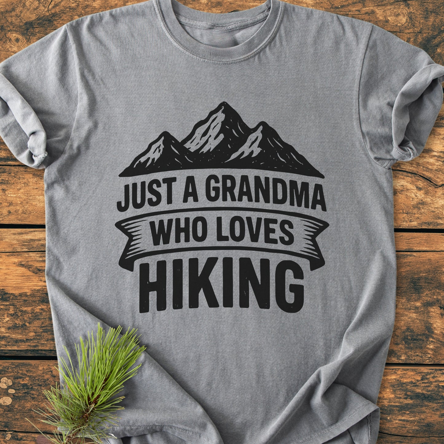 Gray tee featuring Just A Grandma Who Loves Hiking with mountain graphic, presented on wood surface. Pine sprig accentuates the cozy, vintage wash design.