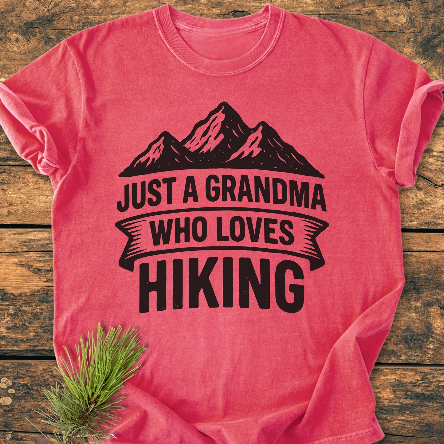 Just A Grandma Who Loves Hiking - Vintage Wash Tee: Vivid red T-shirt with bold black lettering and mountain graphic, styled on a wooden surface with a decorative pine sprig beside it.