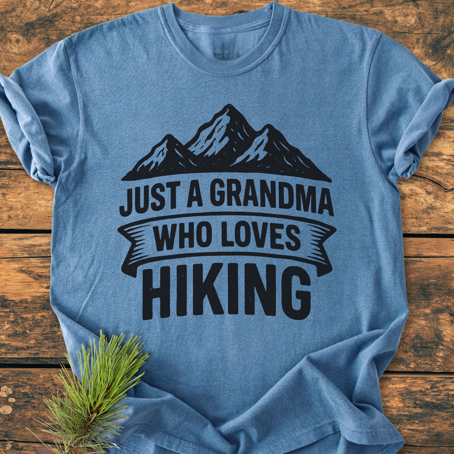Heather-blue Just A Grandma Who Loves Hiking tee featuring mountain silhouette, laid on wood with evergreen accent. Made of soft, 100% ring-spun cotton for comfort and durability.