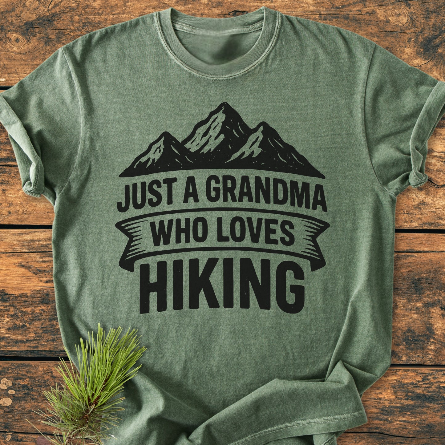 Vintage Wash Tee: A green t-shirt with a mountain silhouette and JUST A GRANDMA WHO LOVES HIKING slogan, laid flat on wood, complemented by a pine sprig, showcasing its relaxed fit and cozy fabric.