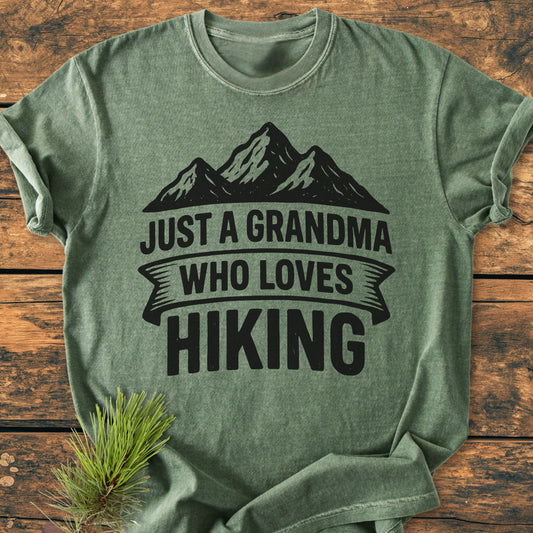 Vintage Wash Tee: A green t-shirt with a mountain silhouette and JUST A GRANDMA WHO LOVES HIKING slogan, laid flat on wood, complemented by a pine sprig, showcasing its relaxed fit and cozy fabric.