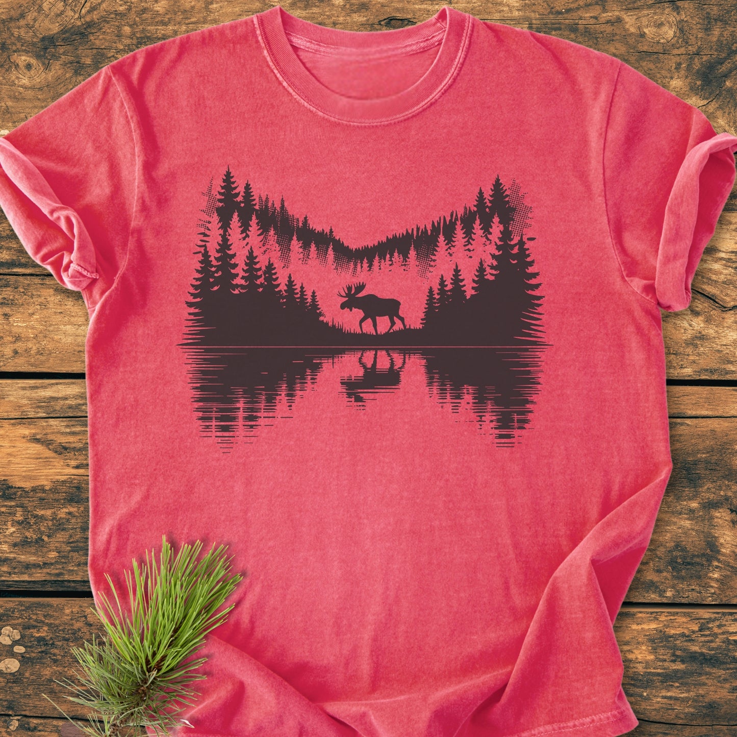 Lone Wanderer - Vintage Wash Tee, featuring a black forest scene with a moose by a lake, displayed on a wooden surface with a pine bough, made from soft, relaxed-fit, garment-dyed cotton.