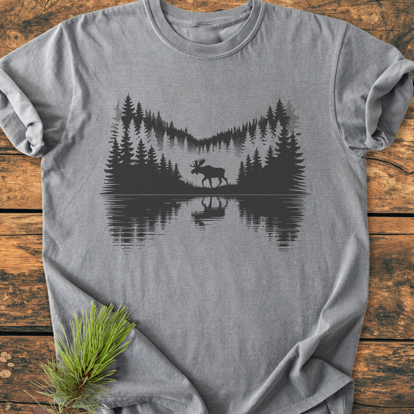 Lone Wanderer - Vintage Wash Tee: Heather-gray shirt with a black forest scene silhouette, featuring a moose by a lake, laid on a wooden surface with a pine sprig beside it.