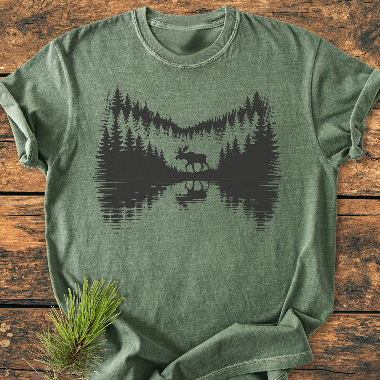 Lone Wanderer - Vintage Wash Tee features a serene lakeshore scene with pine trees and a moose, screen-printed on a heather-green T-shirt, beautifully laid on a rustic wooden surface.