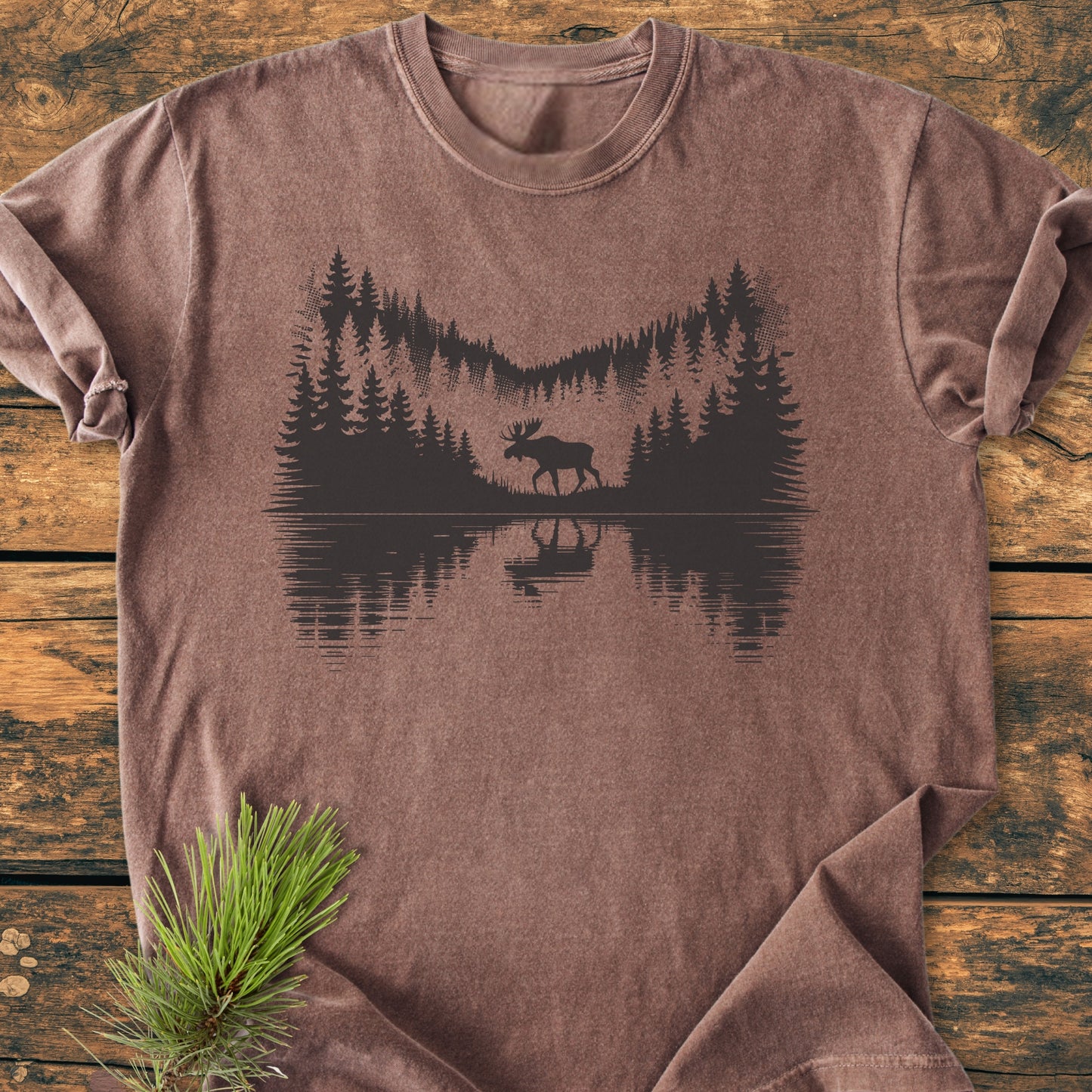 Lone Wanderer - Vintage Wash Tee: brown short-sleeve shirt with a wilderness scene featuring evergreen trees and a moose reflection, laid on a wood-plank surface with a pine sprig accent.