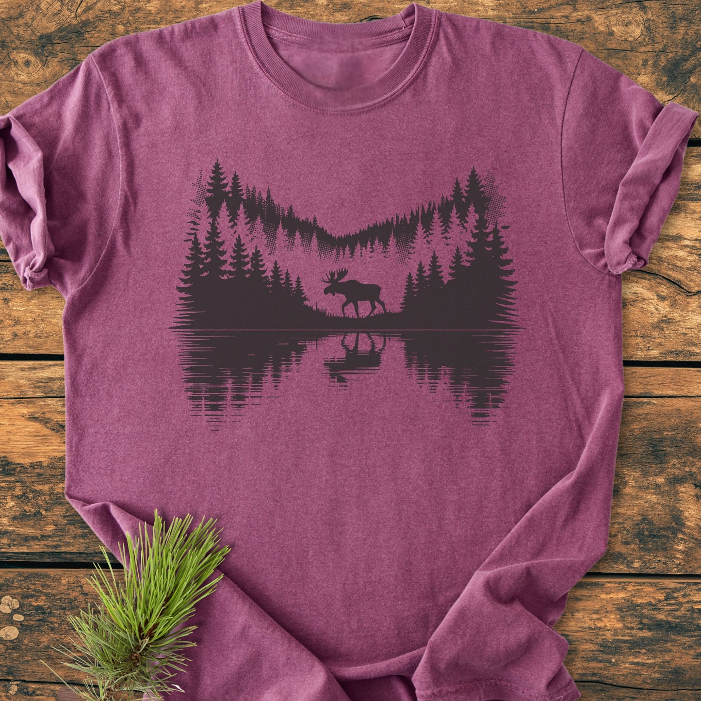 The Lone Wanderer - Vintage Wash Tee features a black pine forest silhouette and moose design on a heathered purple T-shirt, displayed on a rustic wooden surface with a pine sprig beside it.