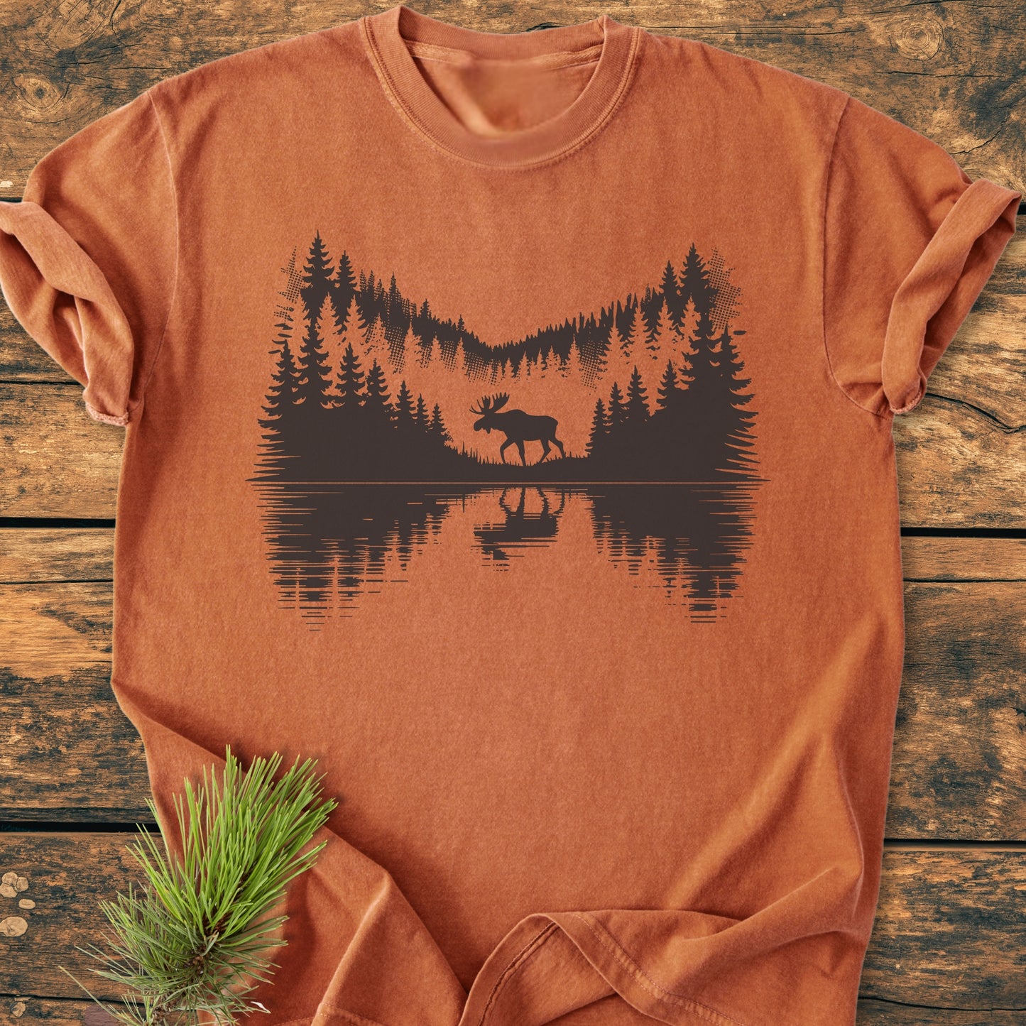Lone Wanderer - Vintage Wash Tee: Rust-orange cotton tee featuring a black wilderness scene with trees and a moose by a lake. Laid on wood surface with pine sprig accent.