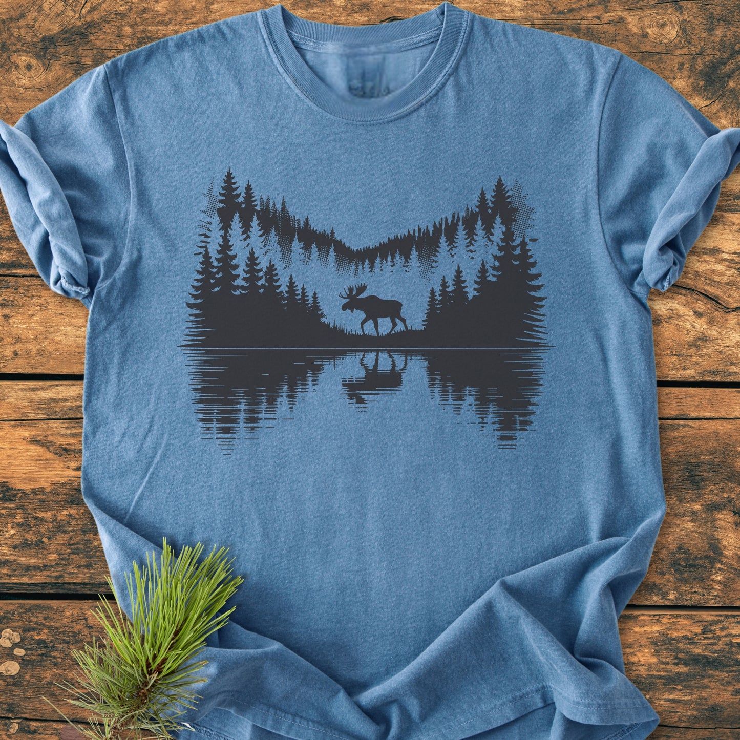 Lone Wanderer - Vintage Wash Tee: Heather-blue T-shirt featuring a dark silhouette of pine trees and a moose by water, laid on a wooden surface, with a pine sprig beside it.