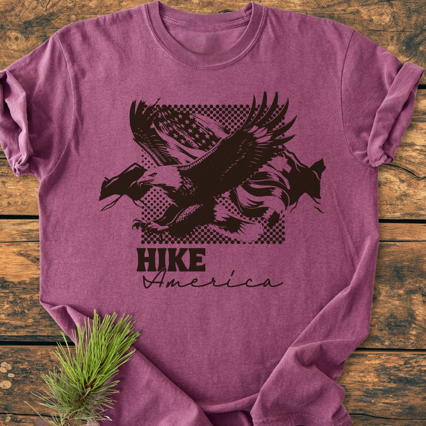 Heather-purple Hike America - Vintage Wash Tee with a stylized bald eagle and mountains design, laid on wood. Soft-washed, relaxed fit, garment-dyed, highlighting its durable, pre-shrunk, ring-spun cotton material.