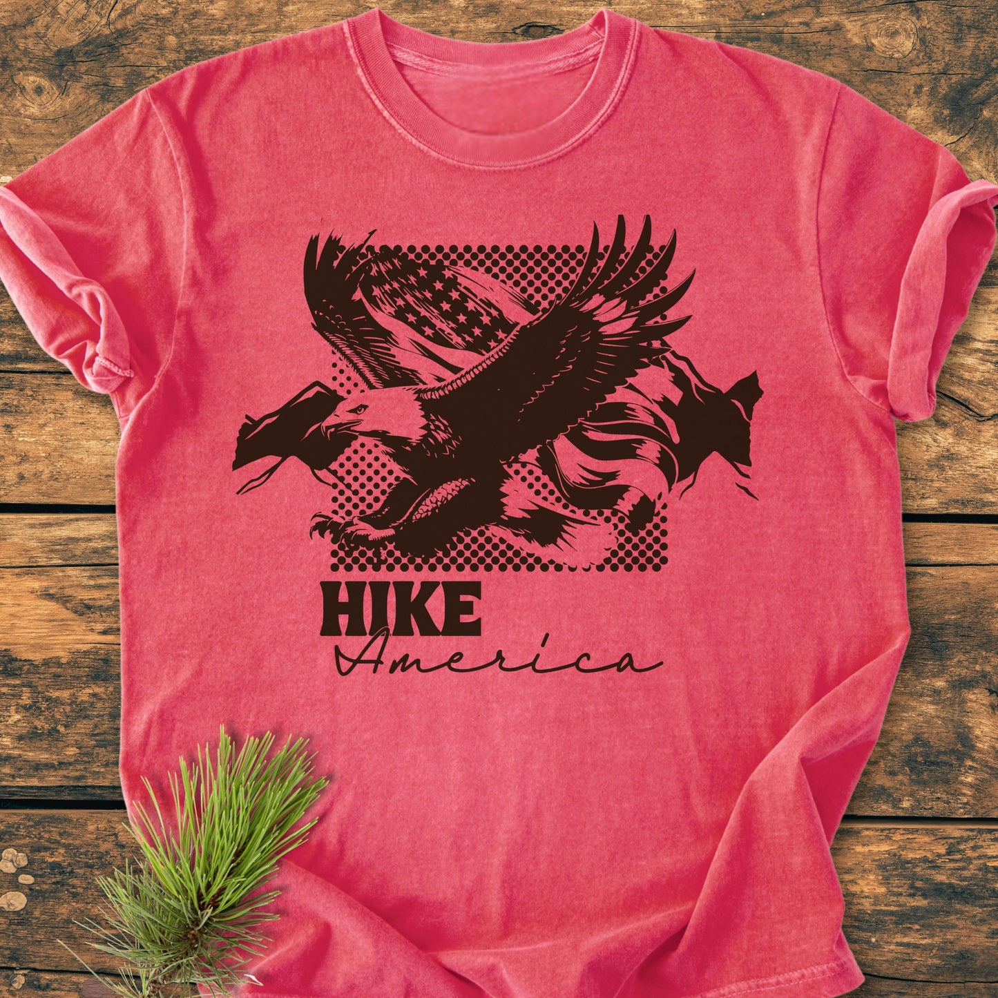 Hike America - Vintage Wash Tee featuring a bold black eagle graphic against an American flag backdrop, laid on wood with a pine sprig, highlighting a relaxed fit and cozy garment-dyed fabric.