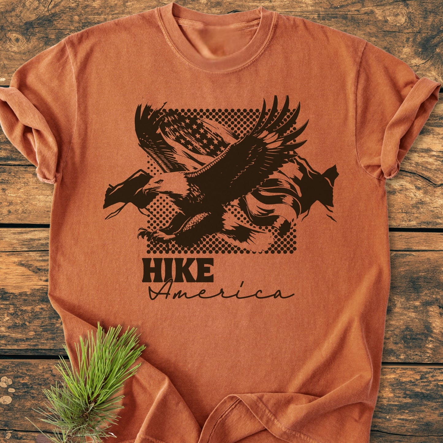 Hike America - Vintage Wash Tee features a bald eagle over a distressed flag motif on a rust-orange T-shirt, laid on wood with a pine sprig, emphasizing a rustic, outdoorsy theme.
