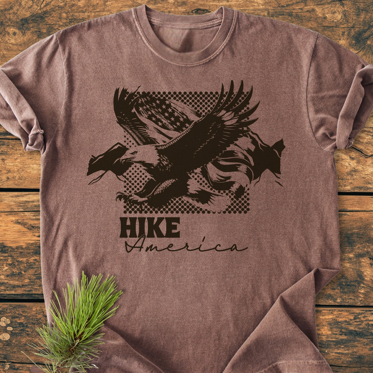 Hike America - Vintage Wash Tee featuring a stylized bald eagle graphic with HIKE America text, laid on wood with a pine bough. Made from soft, durable, pre-shrunk 100% ring-spun cotton.