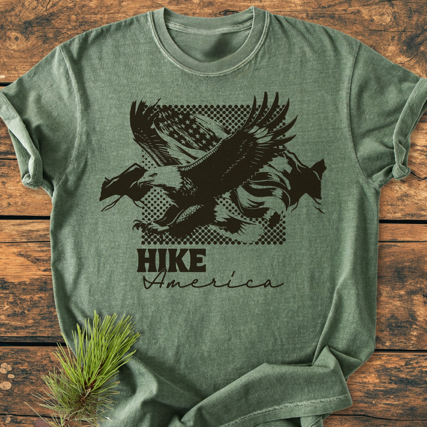 Hike America - Vintage Wash Tee, a green crew-neck shirt with a bold eagle graphic over a halftone flag motif and “HIKE America” text, laid on wooden surface with a pine sprig.