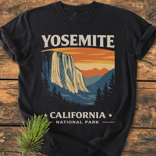 Yosemite National Park - Vintage Wash Tee featuring a stylized sunset design with El Capitan silhouette. Laid flat on wood, showcasing its soft fabric and relaxed fit, with a pine sprig accent.