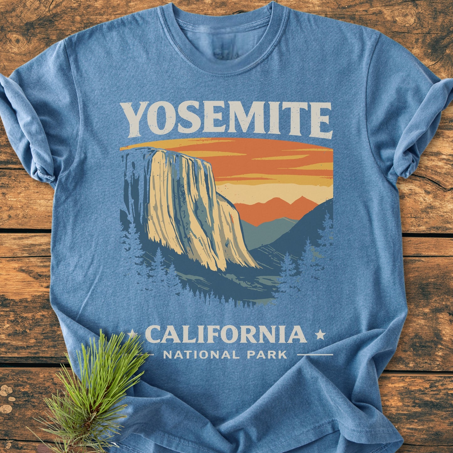 Yosemite National Park - Vintage Wash Tee features a vintage Yosemite design with El Capitan and pine trees. Displayed flat on wood with a pine sprig accentuating its natural appeal.