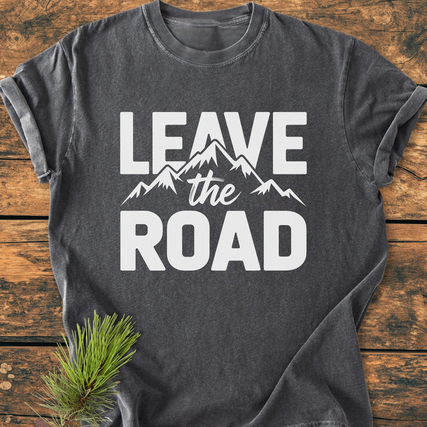 Leave The Road - Vintage Wash Tee in charcoal-gray with bold white text and a mountain graphic, laid flat on a wooden surface beside a pine branch, showcasing its outdoorsy theme.