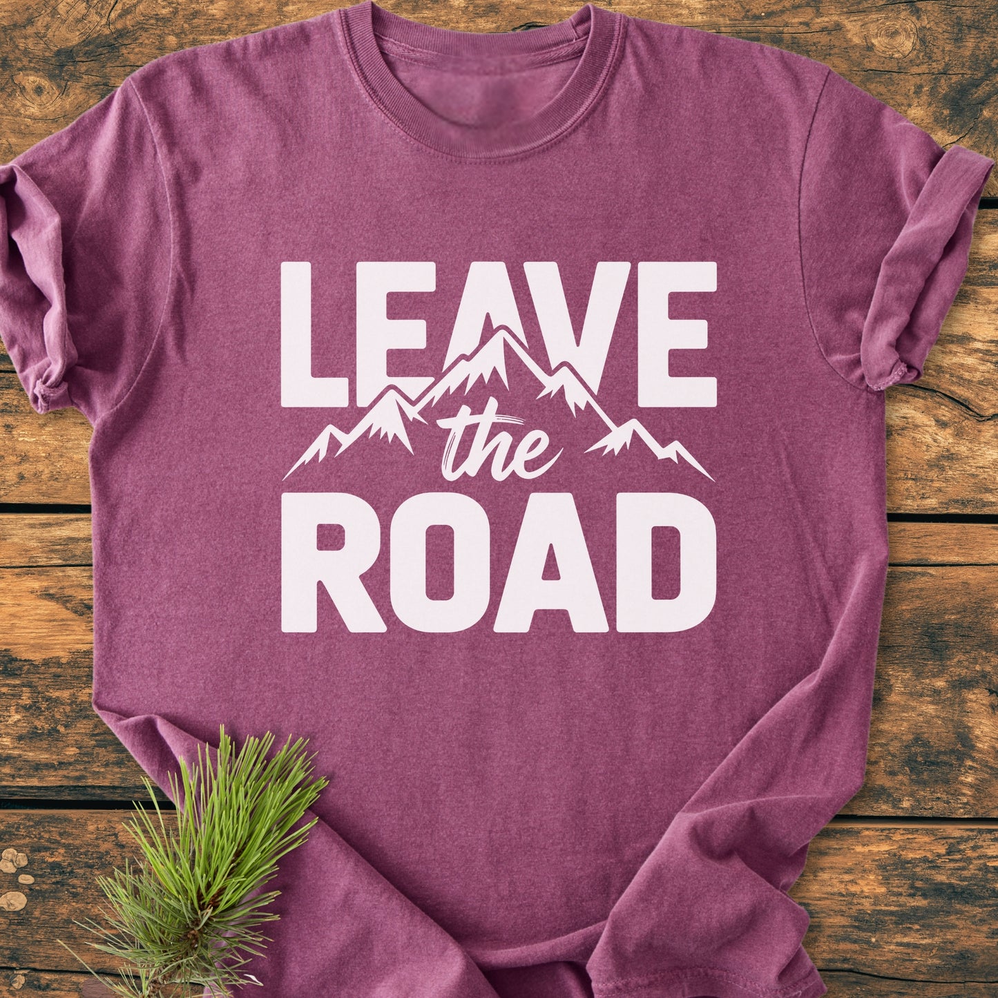 Leave The Road - Vintage Wash Tee: A folded purple-mauve T-shirt with LEAVE the ROAD and mountain graphic in white. Laid on wood, accessorized with a pine sprig, emphasizing an outdoorsy feel.