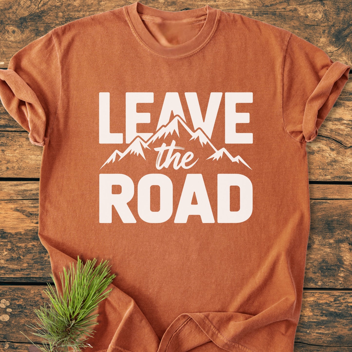 Leave The Road - Vintage Wash Tee displayed on a wooden surface, featuring a bold slogan and mountain graphic. Crafted from soft, garment-dyed cotton, it offers a relaxed fit and durable stitching.