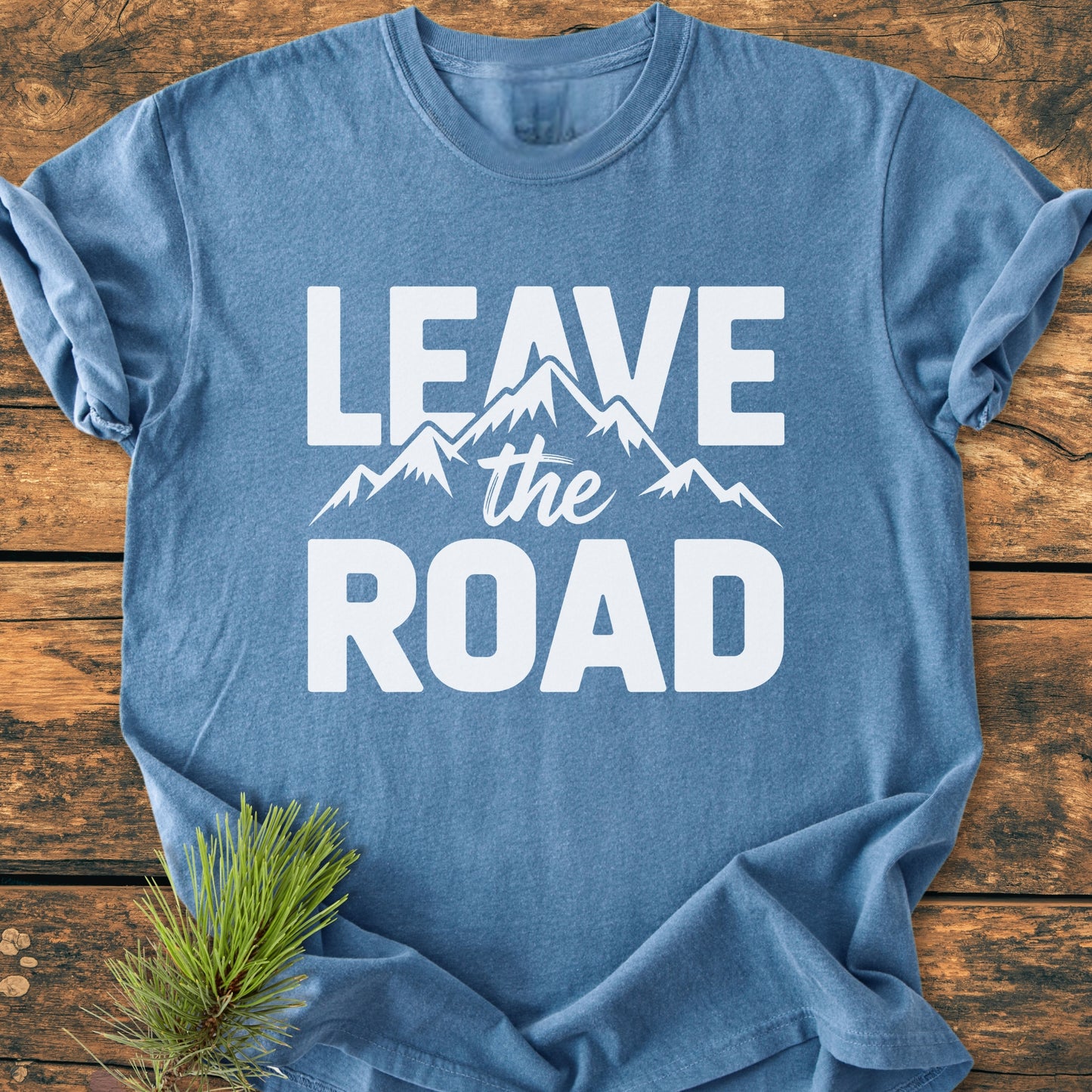 Leave The Road - Vintage Wash Tee, featuring a heather-blue shade with bold white slogan and mountain graphic, laid flat with a pine sprig beside it on a wooden surface.