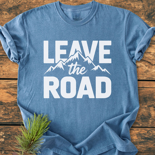 Leave The Road - Vintage Wash Tee, featuring a heather-blue shade with bold white slogan and mountain graphic, laid flat with a pine sprig beside it on a wooden surface.
