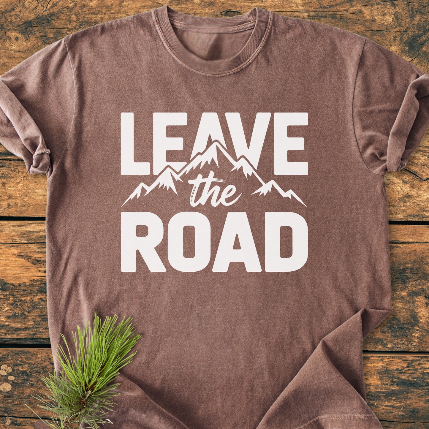 Leave The Road - Vintage Wash Tee featuring a white mountain graphic and bold lettering, laid flat on a wooden surface with a pine sprig accentuating its rustic appeal.