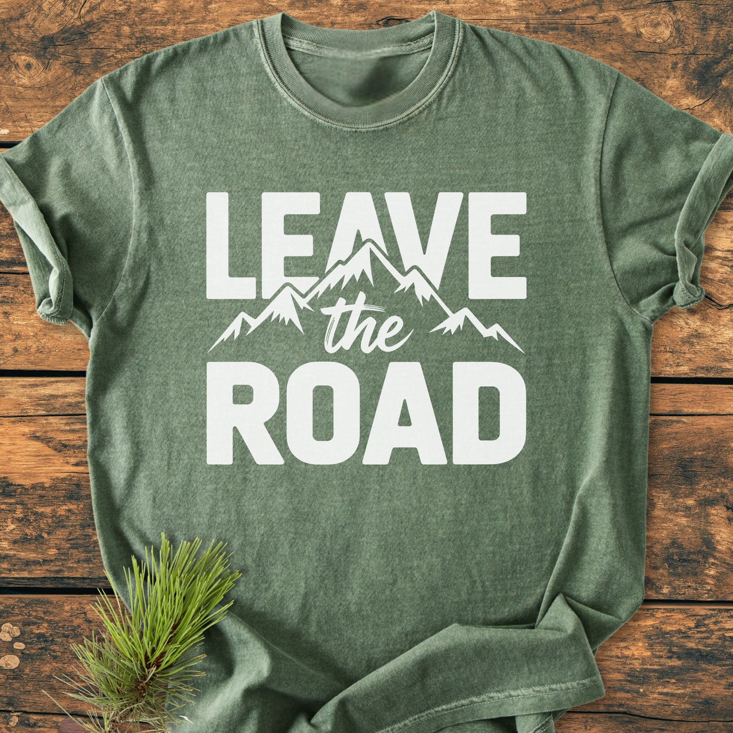 Leave The Road - Vintage Wash Tee displayed on wooden surface, featuring white “LEAVE the ROAD” text and mountain silhouette. A pine branch accents the shirt, emphasizing the outdoors theme.