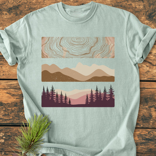 Minimalist Mountain Graphic - Vintage Wash Tee features stacked abstract nature motifs in earth tones on a pale-green surface, emphasizing contour lines, mountain silhouettes, and forest scenes. Displayed on wood with pine sprig.