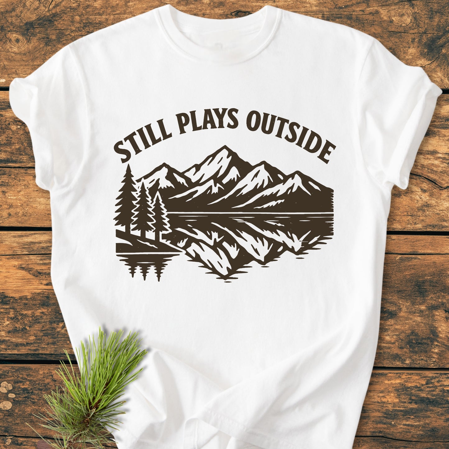 Still Plays Outside - Vintage Wash Tee with a brown outdoors scene of pine trees and mountains, laid on wood. Features soft, garment-dyed 100% ring-spun cotton, relaxed fit, and durable double-needle stitching.