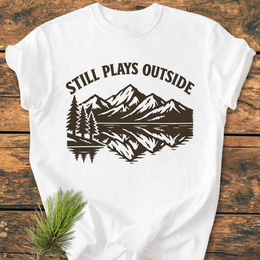 Still Plays Outside - Vintage Wash Tee with a brown outdoors scene of pine trees and mountains, laid on wood. Features soft, garment-dyed 100% ring-spun cotton, relaxed fit, and durable double-needle stitching.