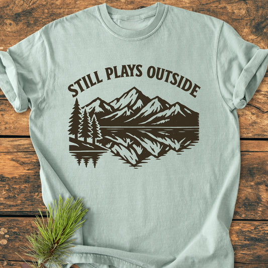 Still Plays Outside - Vintage Wash Tee featuring a dark brown outdoor graphic of pine trees and mountains on a pale-green backdrop, laid on wood with a pine sprig nearby.