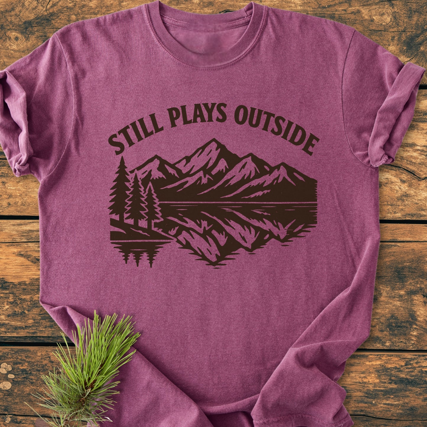 Still Plays Outside - Vintage Wash Tee featuring a mountain scene with pine trees on a heathered purple fabric, laid flat on wood, with a sprig of evergreen beside it.