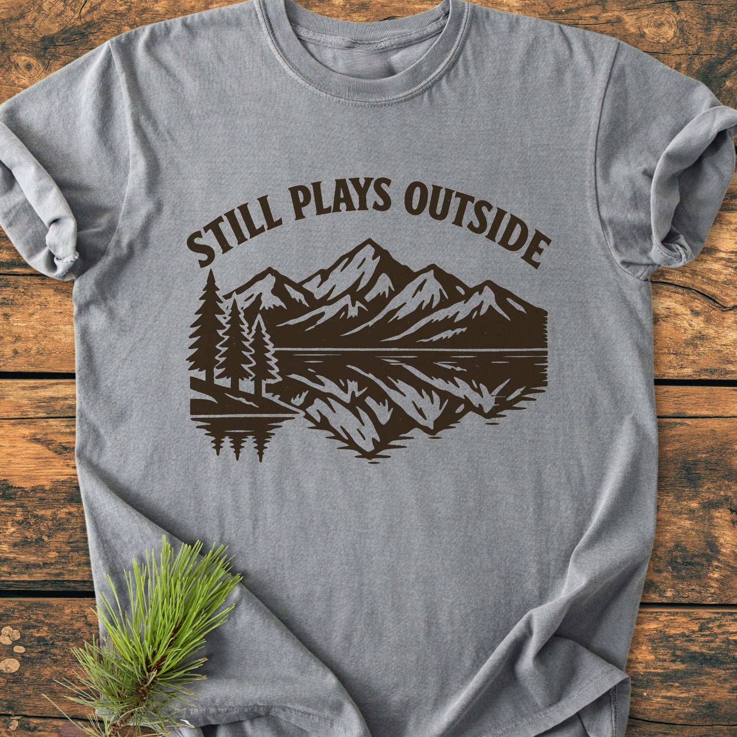 Heather-gray Still Plays Outside - Vintage Wash Tee features a nature scene with pine trees, mountains, and a lake reflection, laid flat on wood with pine needles beside it, highlighting its outdoor theme.