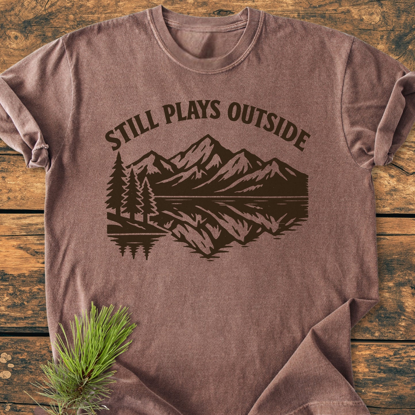 Still Plays Outside - Vintage Wash Tee laid flat, featuring a stylized outdoor scene with mountains and trees. Made from soft, garment-dyed, ring-spun cotton, offering a relaxed fit.