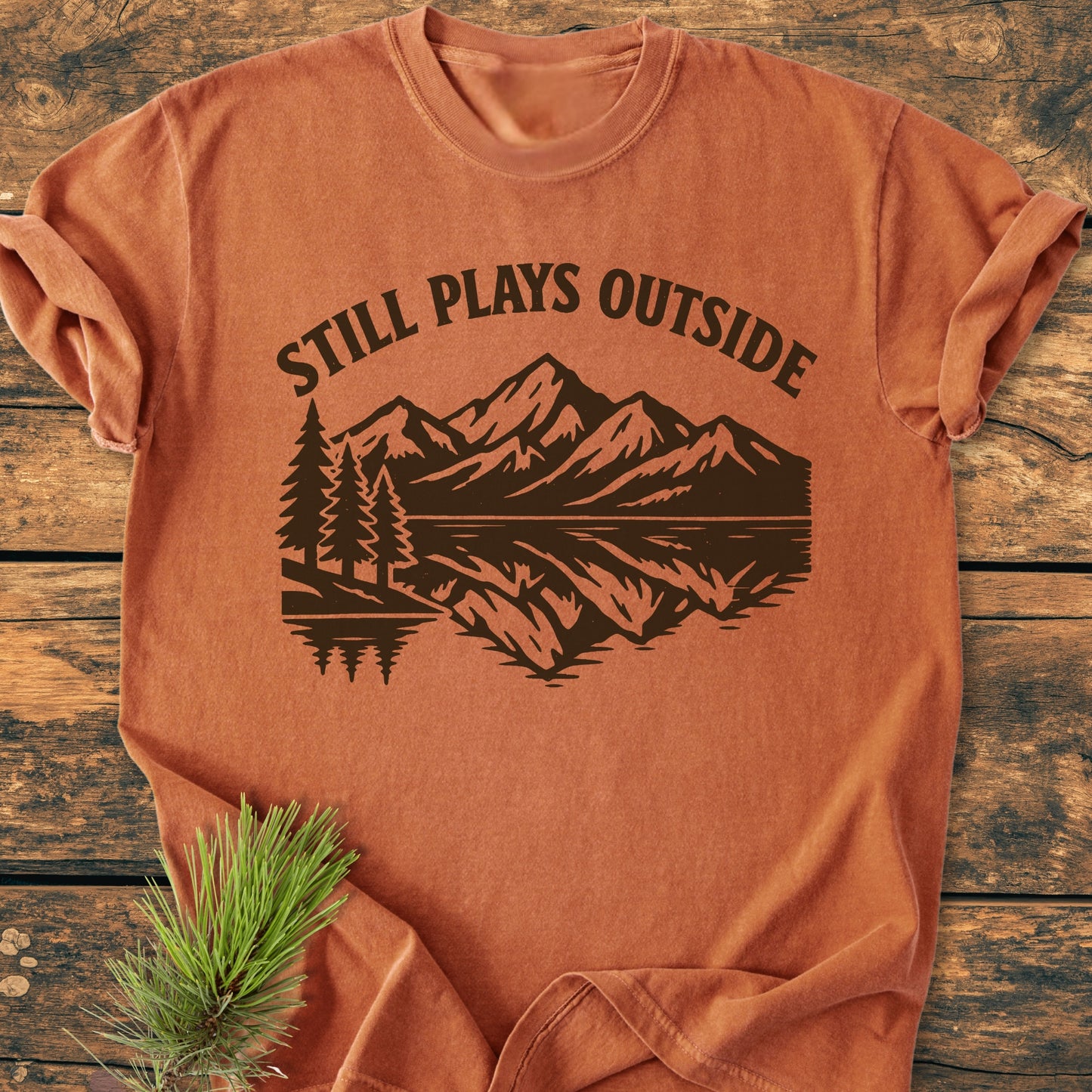 Still Plays Outside - Vintage Wash Tee featuring a mountain and pine tree silhouette with a lake reflection, styled with a pine sprig, on a rustic wooden background, highlighting its casual, durable design.