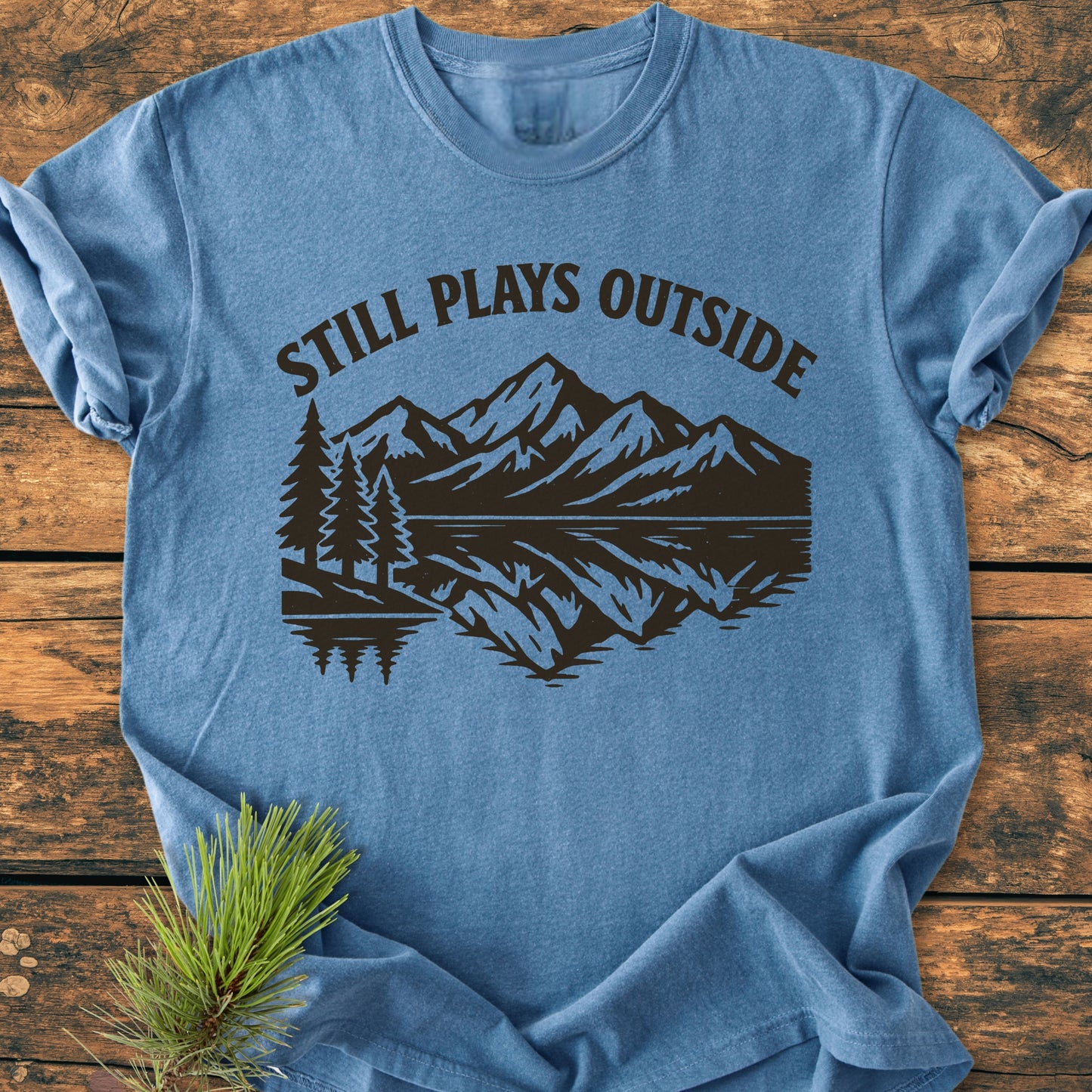Still Plays Outside - Vintage Wash Tee features a stylized mountain-and-lake scene on a heather-blue fabric, emphasizing its relaxed fit and soft, garment-dyed finish, displayed on a wooden surface with pine accents.