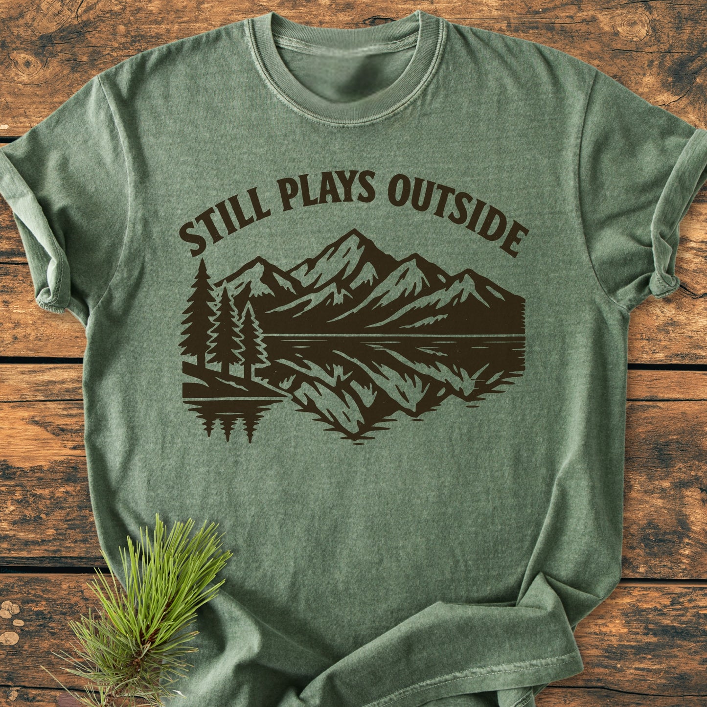 Still Plays Outside - Vintage Wash Tee features a mountain range and lake graphic on heather-green fabric, laid on wood with pine needles, showcasing its relaxed fit and durable, soft-washed cotton design.